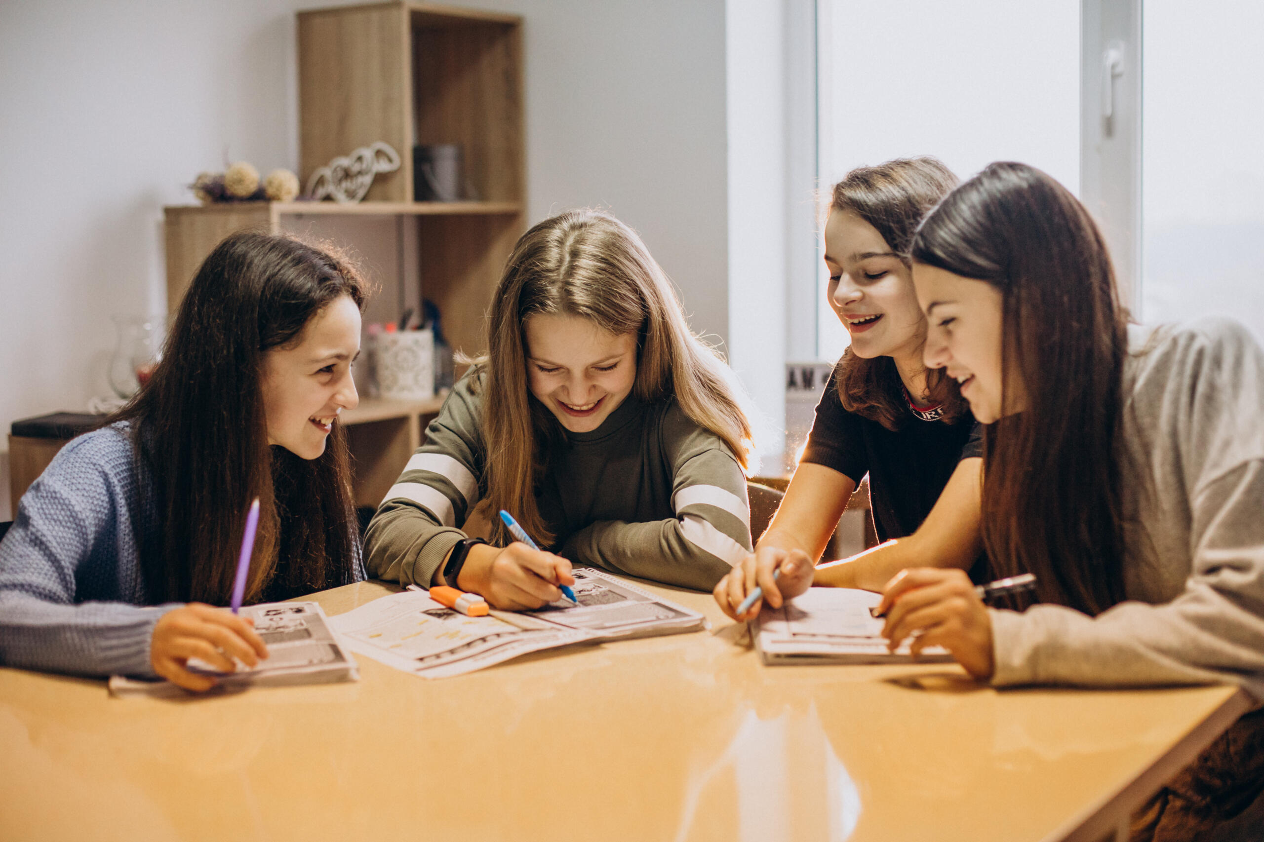 Four girls sit around a table, smiling as they write and draw in open notebooks with colored pens.
